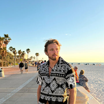 Man wearing a black and white patterned shirt standing on a promenade with palm trees and people in the background.